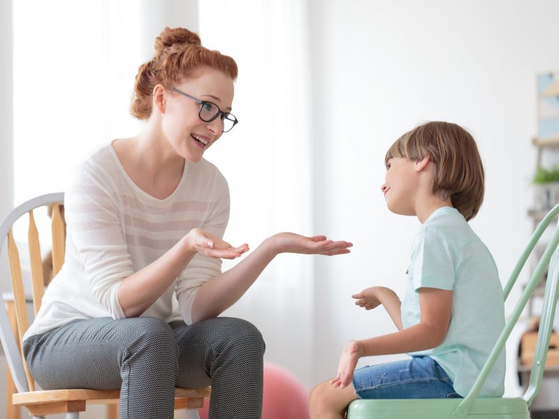 Smiling psychotherapist talking with autistic boy sitting on mint chair during psychotherapy